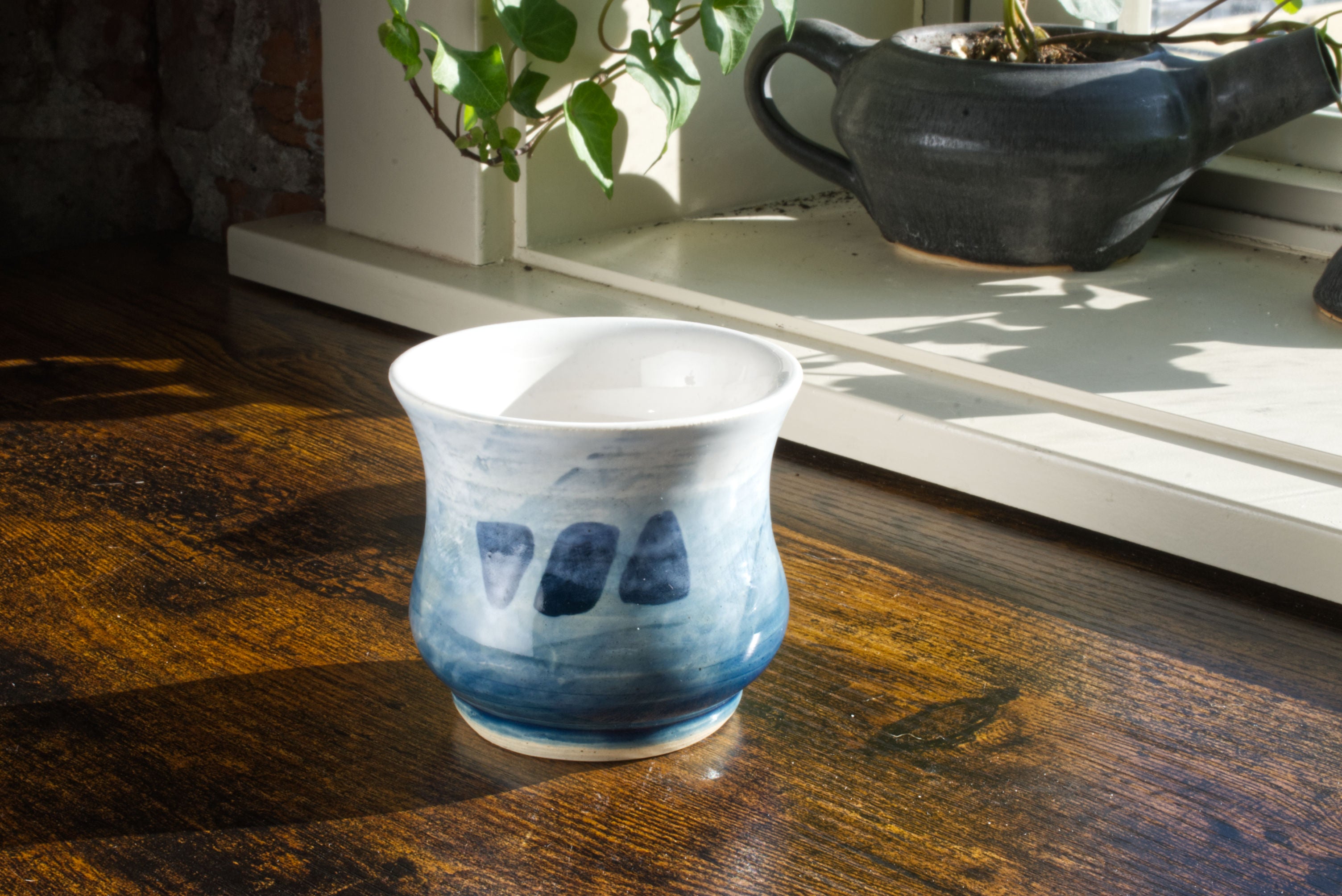 Handmade handleless coffee mug, glazed in a blue to white gradient to look like the ocean, with the brekky threads logo hand painted on the front. The curved sides of the mug are smooth to represent the calming aesthetic of the bay. Seen from the front, on a desk, with a teapot planter in the background.