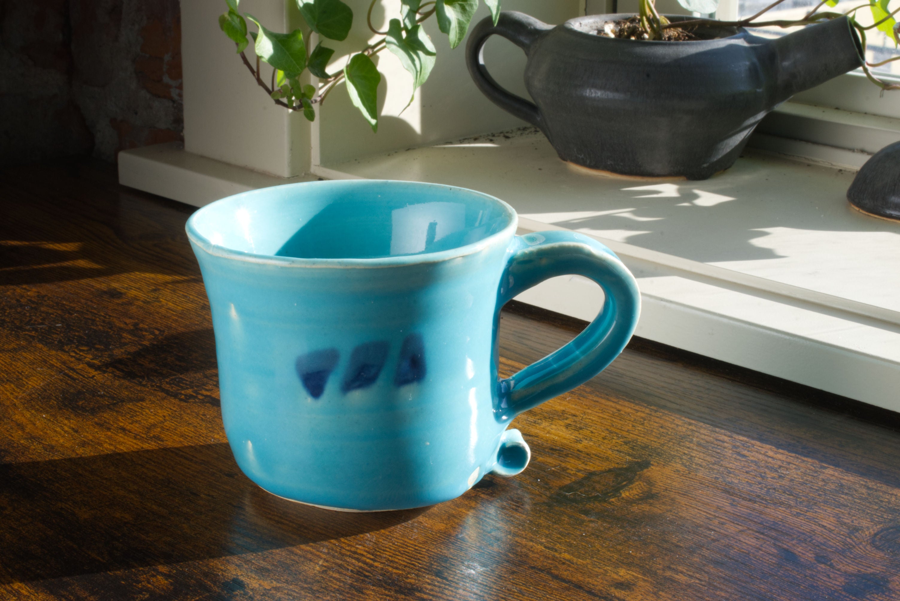 Handmade stoneware mug with curved handle and hand painted Brekky Threads Logo on the side. The entire mug is glazed in blue poppy, which is a bright and vibrant sky blue, while the logo is a deep navy blue shining through from underneath the blue poppy. Pictured on a wooden desk with a handmade teapot planter in the background. Color: Blue Poppy. Unisex.