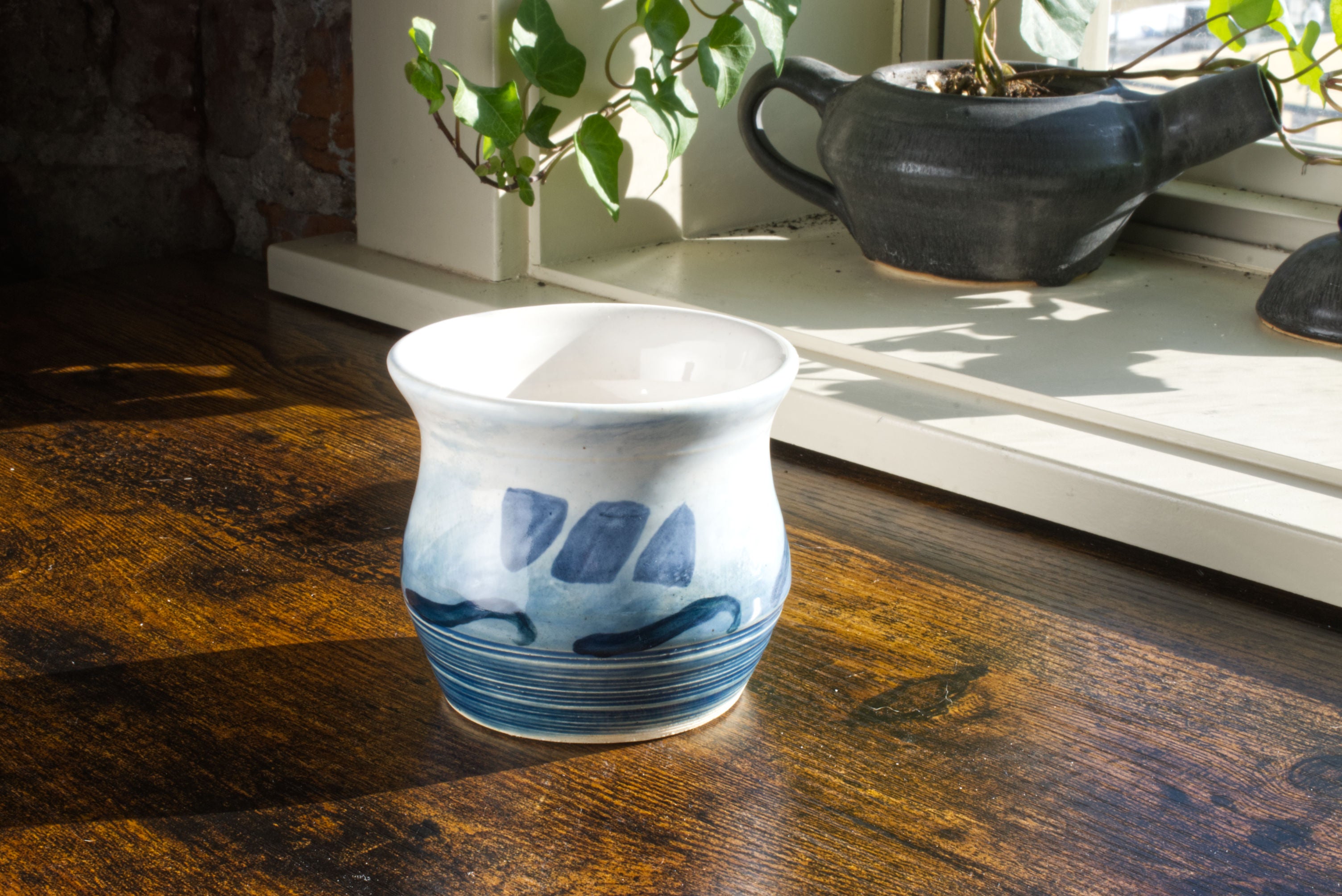 Handmade handleless coffee mug, glazed in a blue to white gradient to look like the ocean, with the brekky threads logo hand painted on the front, with waves painted along the middle. The foot of the mug has rough texture to mimic the roughness of the ocean. Seen from the front, on a desk, with a teapot planter in the background.