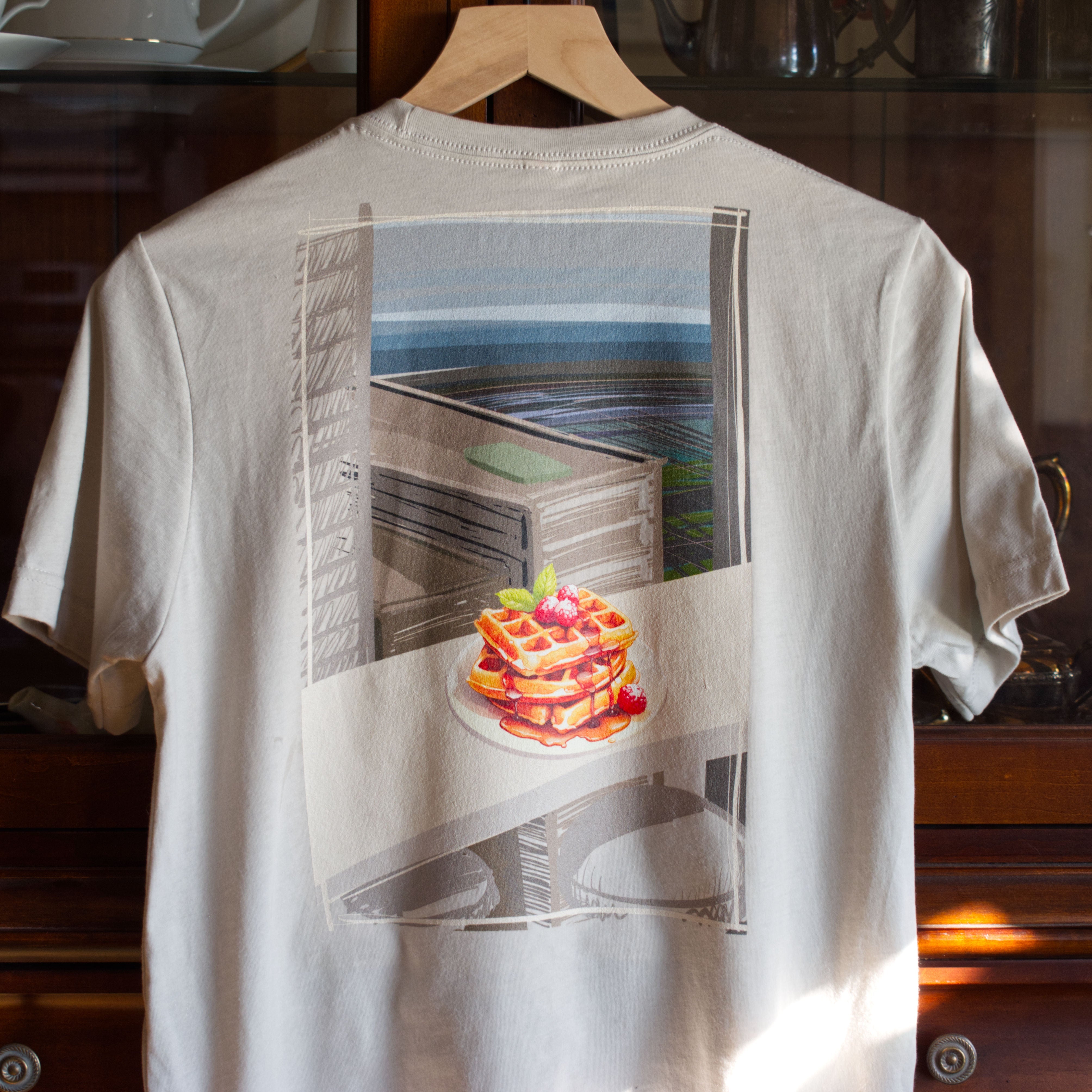 White t-shirt with a hand drawn graphic design: waffles sitting on a countertop in a sky-rise, looking out over a city. The t-shirt is called Waffle city by Brekky Threads. Color: Heather Dust.