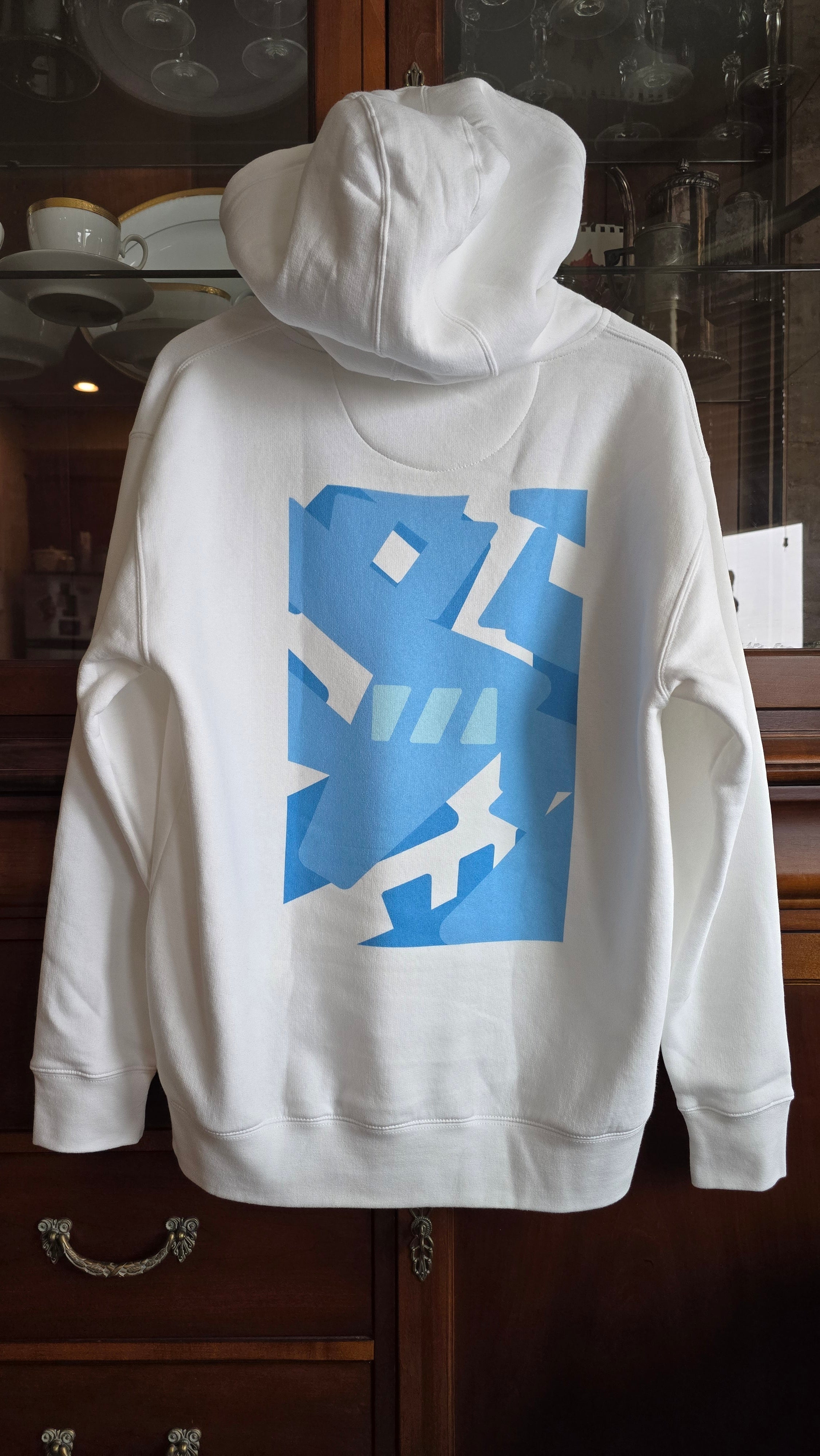White hoodie hanging on a coat hangar, with the back facing the camera. The River of Life is printed in bright blue on its back.