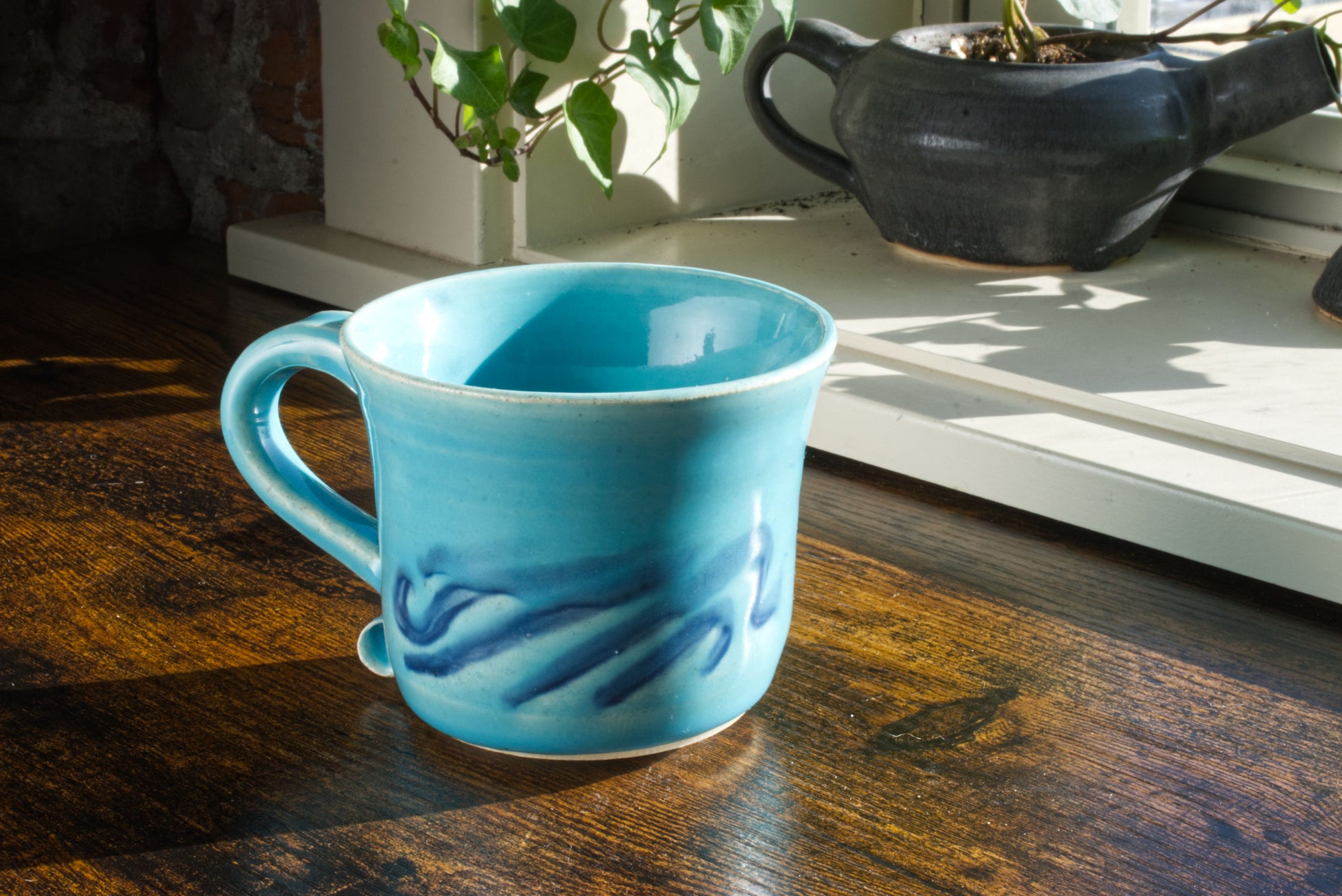 Handmade stoneware mug with curved handle and hand painted waves on the side, on the lower half. The waves themselves can be felt as a relief due to the way the glaze flowed. The entire mug is glazed in blue poppy, which is a bright and vibrant sky blue, while the waves are a deep navy blue shining through from underneath the blue poppy. Pictured on a wooden desk with a handmade teapot planter in the background. Color: Blue Poppy. Unisex.