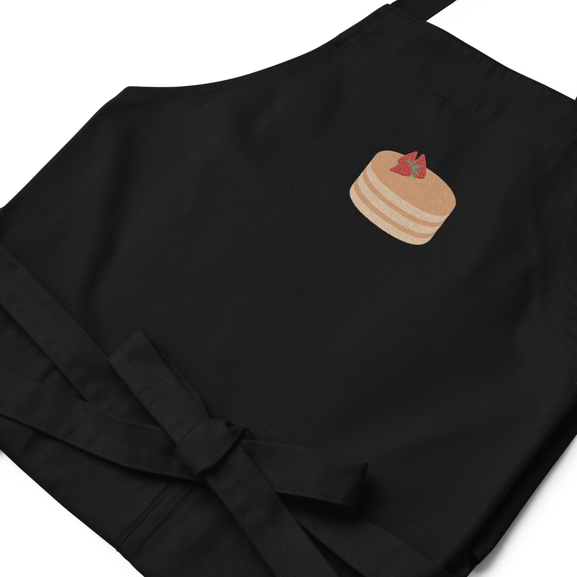 Closeup of kitchen apron embroidered on the front. The design is of pancakes with 3 strawberries on top, in a graphic are / vector art style. Apron features 2 front pockets and long straps to be tied behind the wearer in the usual manner. Unisex. One Size. Color: Black. Designed by Brekky Threads.