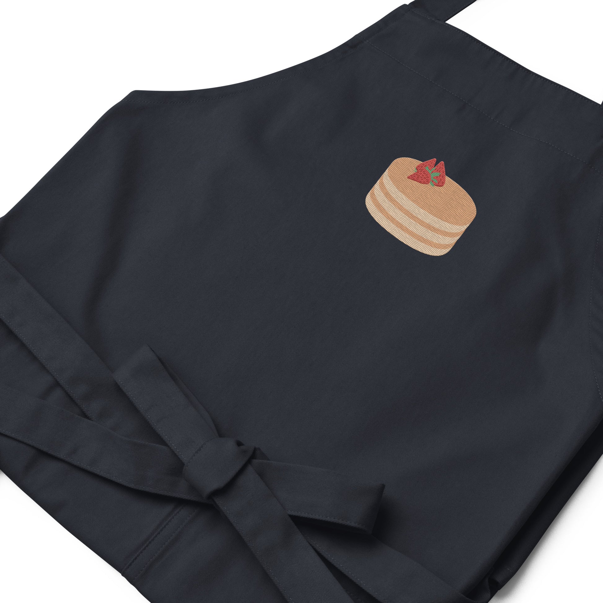 Closeup of kitchen apron embroidered on the front. The design is of pancakes with 3 strawberries on top, in a graphic are / vector art style. Apron features 2 front pockets and long straps to be tied behind the wearer in the usual manner. Unisex. One Size. Color: Navy. Designed by Brekky Threads.