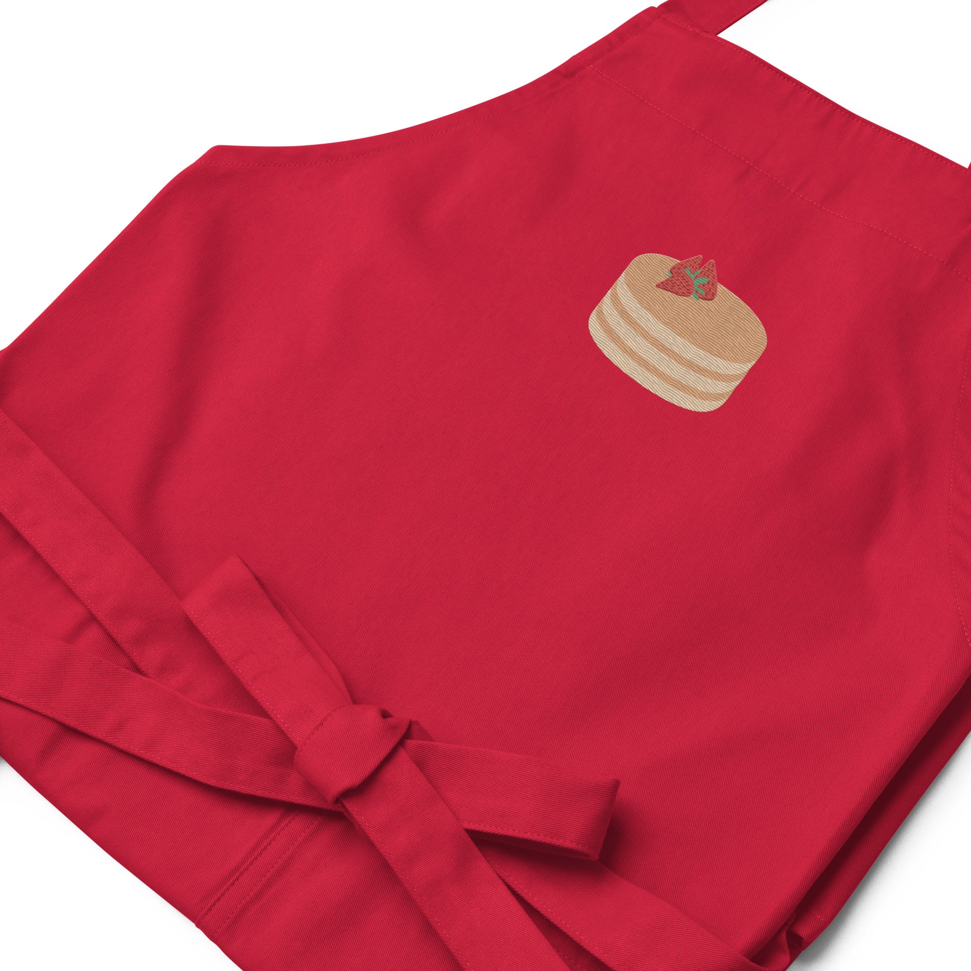Closeup of kitchen apron embroidered on the front. The design is of pancakes with 3 strawberries on top, in a graphic are / vector art style. Apron features 2 front pockets and long straps to be tied behind the wearer in the usual manner. Unisex. One Size. Color: Red. Designed by Brekky Threads.