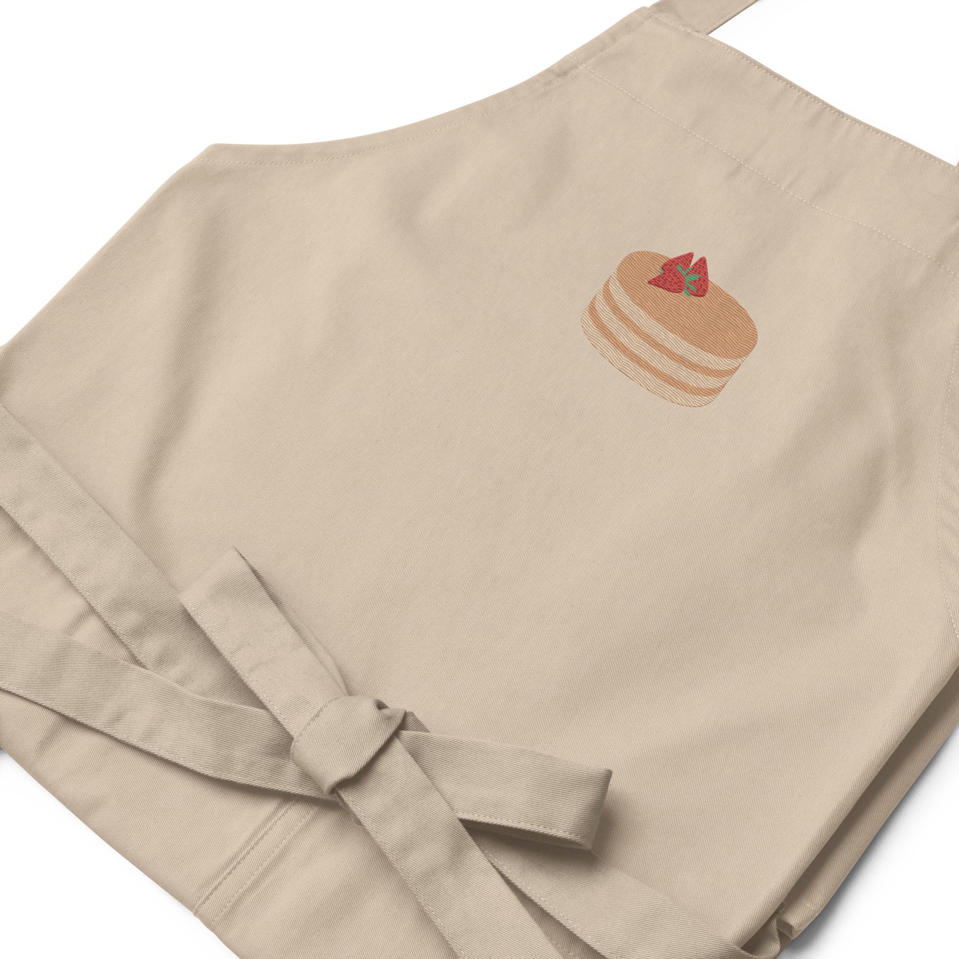 Closeup of kitchen apron embroidered on the front. The design is of pancakes with 3 strawberries on top, in a graphic are / vector art style. Apron features 2 front pockets and long straps to be tied behind the wearer in the usual manner. Unisex. One Size. Color: Rope (Beige). Designed by Brekky Threads.