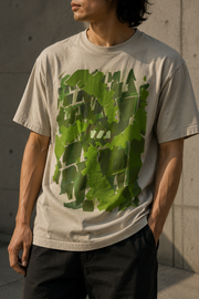 White man standing with one hand in his pocket, wearing an oversized faded bone tshirt with the Emerald Stream design by Brekky Threads on the front. The design is composed of the Brekky logo in different shades of green arranged artistically to form a swirl.