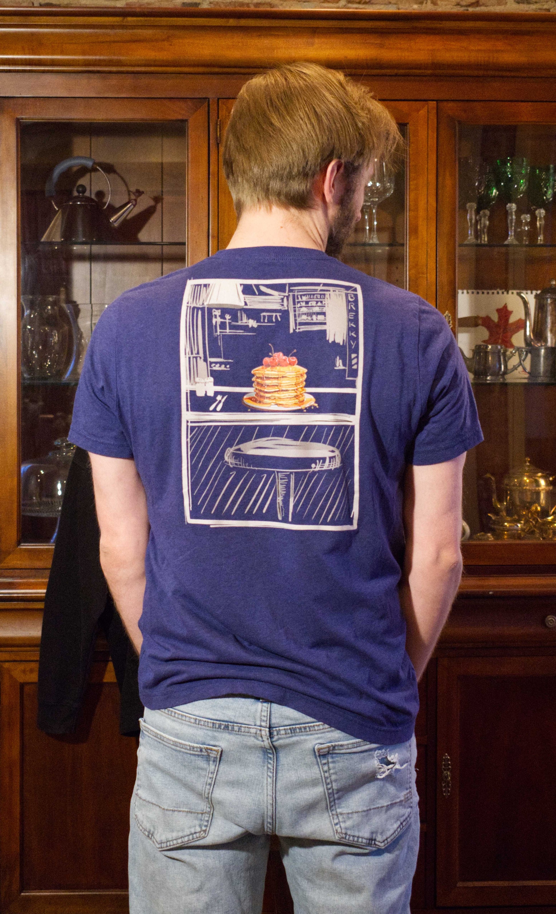 This slim fitting stretchy tshirt features a sketch style design by Brekky Threads. Pancakes on a plate, drawn in full color, set in a line art breakfast diner sketch. The lines are different shades of grey, while the background takes on the color of the tshirt fabric. Unisex. Color:  Heather Midnight Navy. There is an attractive man standing in the frame with his back turned, wearing the pancake diner tshirt
