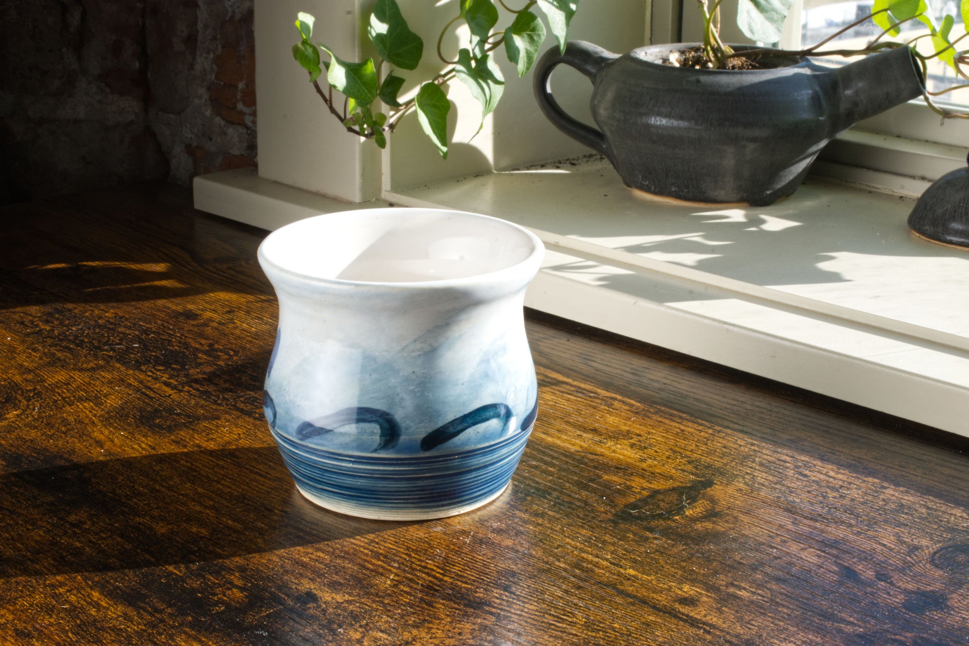 Handmade handleless coffee mug, glazed in a blue to white gradient to look like the ocean,  with waves painted along the middle. The foot of the mug has rough texture to mimic the roughness of the ocean. Seen from the back, on a desk, with a teapot planter in the background.