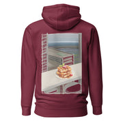 Comfy and cozy hoodie with flat drawstrings. Pictured from the back, a large color print of waffles, plated, on a bar table in front of a window with sky-scrapers, a lower city, and the ocean in the background. Unisex. Color: Maroon.