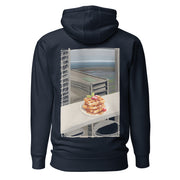Comfy and cozy hoodie with flat drawstrings. Pictured from the back, a large color print of waffles, plated, on a bar table in front of a window with sky-scrapers, a lower city, and the ocean in the background. Unisex. Color: Navy Blazer.